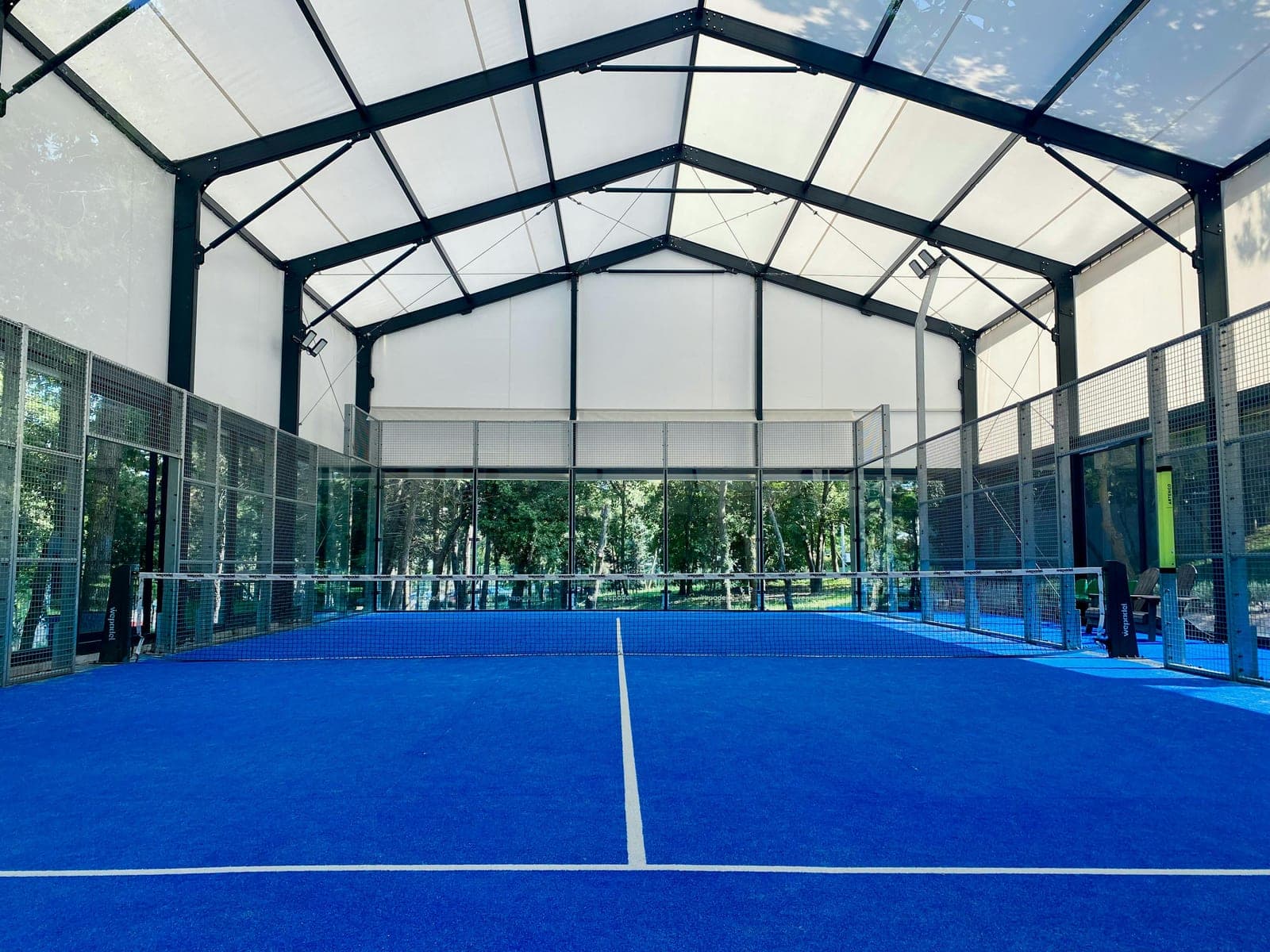 Empty blue padel court inside a translucent canopy structure with glass walls and woodland visible behind